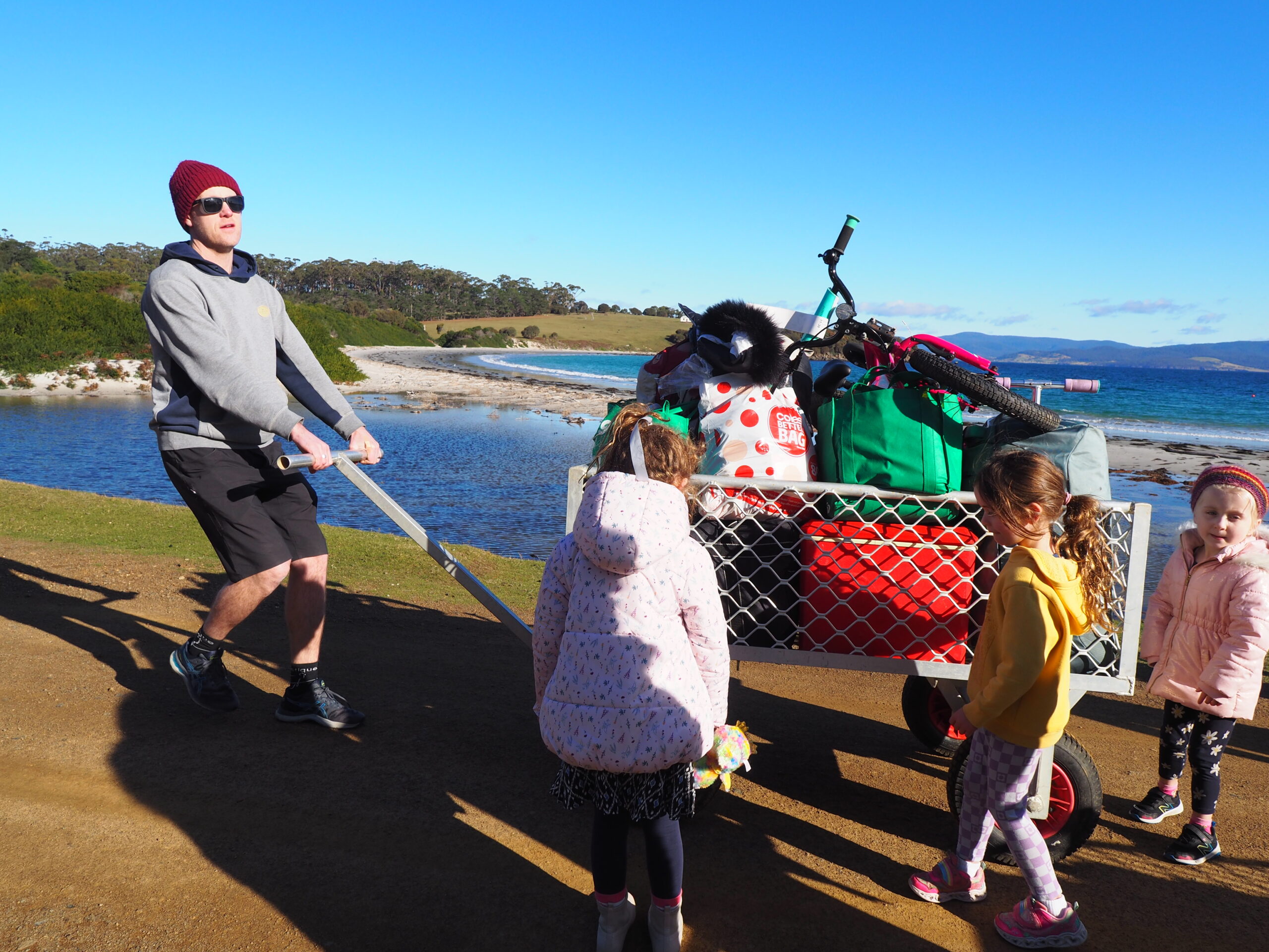 Picture of man hauling a caged trolley with luggage near the sea. Also shows three children