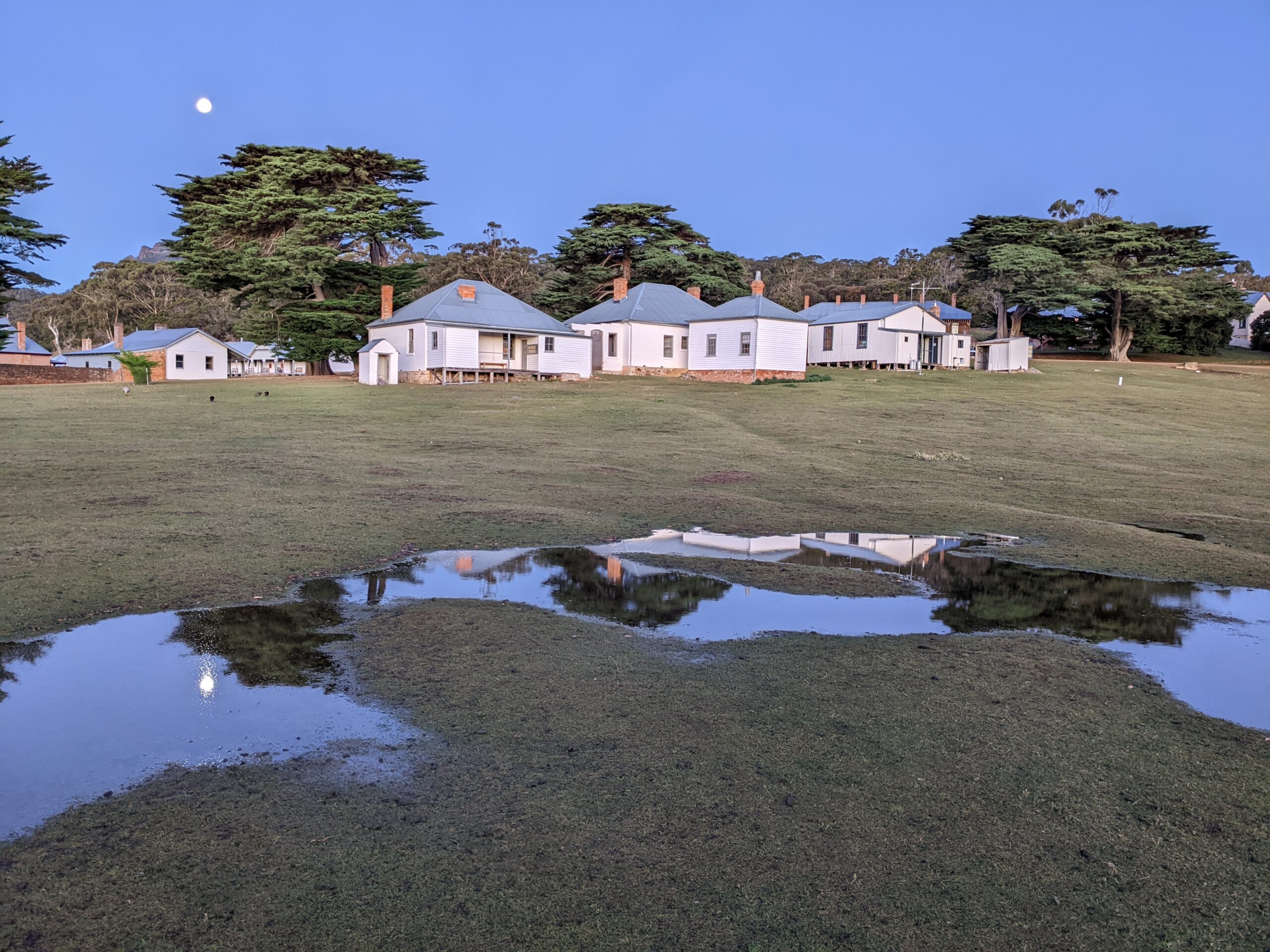 White-washed officer's buildings with a rising moon in the background. Reflections in pools of rainwater in the foreground.er in the foreground.